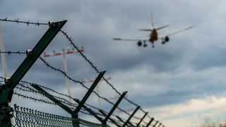 Ein Flugzeug befindet sich im Landeanflug auf den Rheinmain-Airport, im Vordergrund ein Zaun mit Stacheldraht.