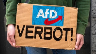 Eine Person hält ein Pappschild mit der Aufschrift "AfD-Verbot!" in den Händen.