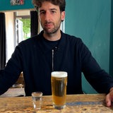 Ein Mann sitzt an einem Tresen vor einem Glas Bier und einem kleinen Schnapsglas mit farblosem Inhalt.