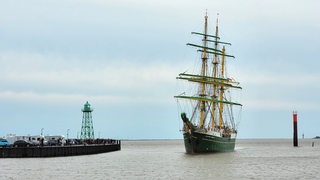 Menschen beobachten von einer Kaimauer aus ein vorbeifahrendes Segelschiff.