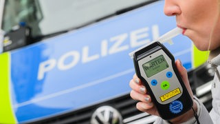 Eine Frau pustet in ein Atemalkoholtestgerät, im Hintergrund steht ein Polizeiwagen.