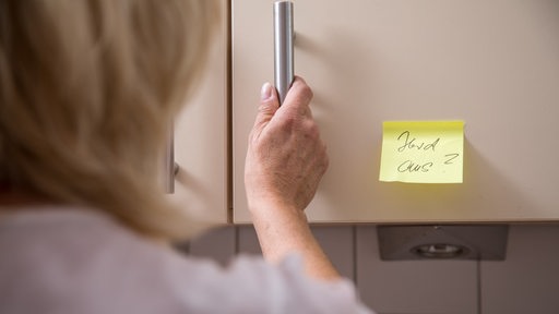 Eine alzheimerkranke Frau öffnet einen Küchenschrank, auf dem ein Hinweis "Herd aus?" steht (Symbolbild)