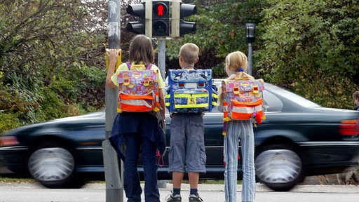 Drei Grundschüler mit Ranzen stehen an einer Fußgängerampel.