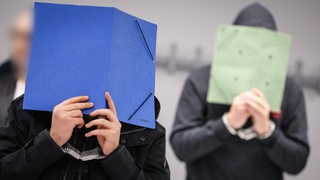 Zwei Angeklagte halten sich im externen Sitzungssaal des Landgerichts Bremen Mappen vor die Gesichter.