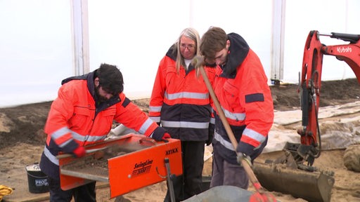 Zwei Mitarbeiter und eine Mitarbeiterin der Landesarchäologie Bremen stehen in einem Zelt und sieben mit einem Gerät Erde.