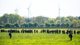 Dutzende Menschen stehen in einer Kette auf einer Wiese. Im Hintergrund sind Bäume und Windräder zu sehen.