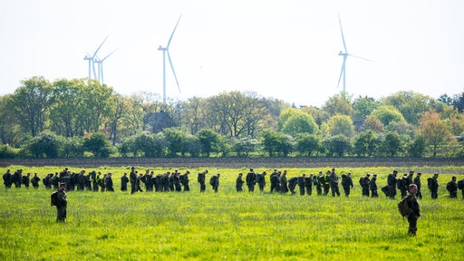 Dutzende Menschen stehen in einer Kette auf einer Wiese. Im Hintergrund sind Bäume und Windräder zu sehen.