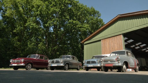 Es stehen mehrere Oldtimer vor einer großen Garage.