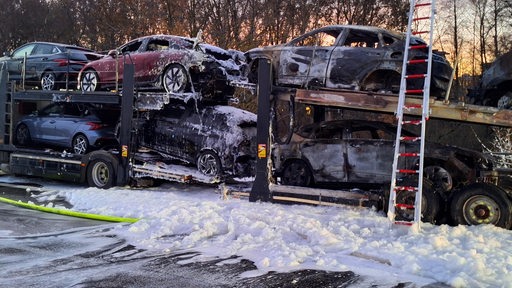 Ein ausgebrannter Autotransporter steht auf einer Autobahn. Weißer Löschschaum liegt auf dne Autos und auf der Fahrbahn.