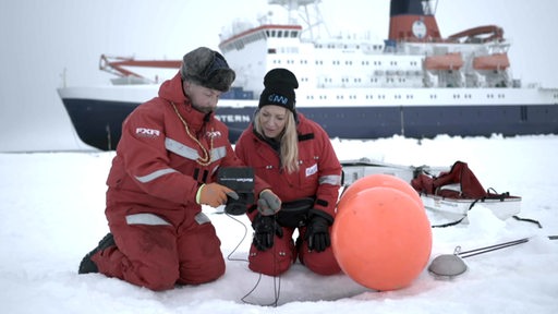 AWI Chefin Antje Boetius und ein Kollege blicken auf ein Tablet, im Hintergrund das Forschungsschiff Polarstern