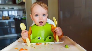 Ein Baby sitzt auf einem Hochstuhl und hält einen Löffel und eine Gabel in der Hand.