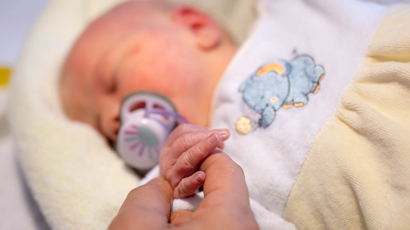 Ein neugeborenes Baby mit einem Schnuller im Mund hält eine Hand