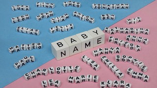 Vier Würfel in der Mitte des Bildes zeigen das Wort "BABY NAME", rundherum liegen viele weitere kleinere Würfel, die andere Namen zeigen.