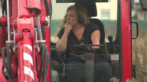 Eine Frau in einem Bagger. Sie schaut nachdenklich zur Seite.