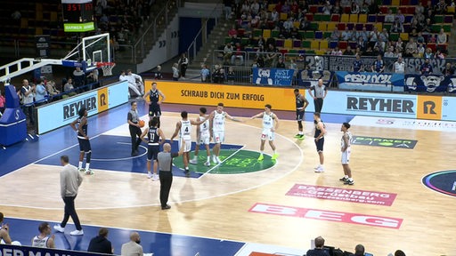 Auf einem Basketballfeld stehen mehrere Spieler zweier Mannschaften über dem linken Spielfeld verteilt