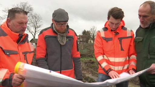 Vier Männer mit orangenen Warnjacken gucken sich gemeinsam einen Bauplan an.