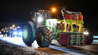 Ein Traktor transportiert das schwarz-rot-goldene Modell einer Kuh und steht gemeinsam mit anderen Traktoren auf einer Autobahn, die blockiert ist.