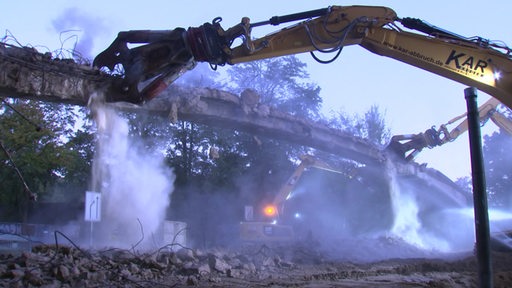 Mehrere Baggerarme zerstören die Überreste einer abgerissenen Fußgängerbrücke