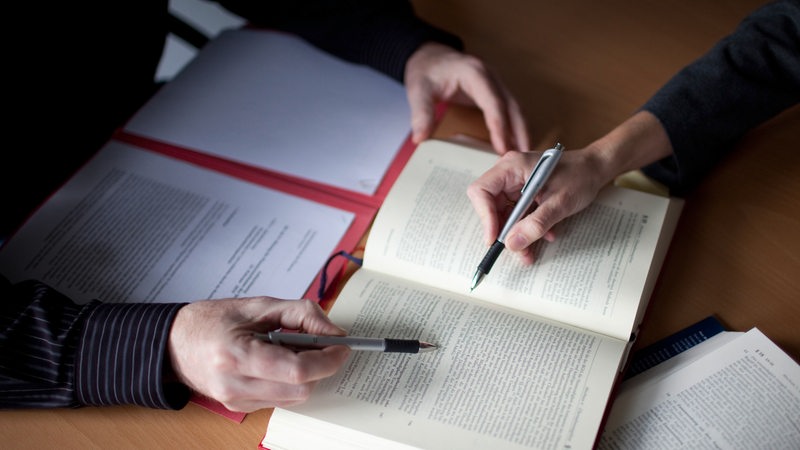 Menschen sitzen an einem Tisch mit aufgeschlagenen Büchern und zeigen mit Stiften aus Passagen (Symbolbild)