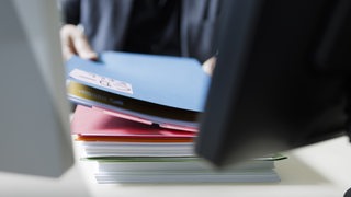 Ein Mann legt mehrere Akten zwischen zwei Computer-Bildschirmen auf einen Schreibtisch.