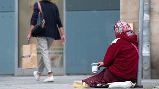 Eine Frau mit Kopftuch bettelt mit einem Pappbecher um Geld. Im Hintergrund eine Frau mit Shopping-Tüte.