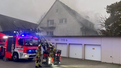 Zwei Feuerwehrleute stehen vor einem Einsatzfahrzeug. Im Hintergrund ist ein brennendes Haus zu sehen.