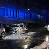 Vor einem Wohnhaus in Bremen-Gröpelingen stehen ein ausgebranntes Auto und ein Feuerwehr-Rettungswagen mit Blaulicht.