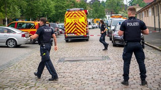 Drei Polizisten riegeln eine Straße ab, in der ein Wagen der Feuerwehr steht.