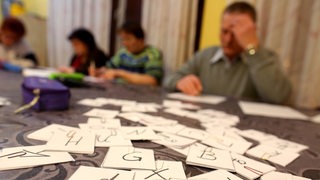 Auf einem Tisch liegen mehrere Karten mit Buchstaben, im Hintergrund schreiben Erwachsene auf Zettel.