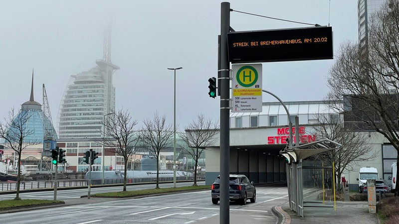 Vor einer leeren Bushaltestelle leuchtet ein Hinweis auf einer Anzeigetafel.