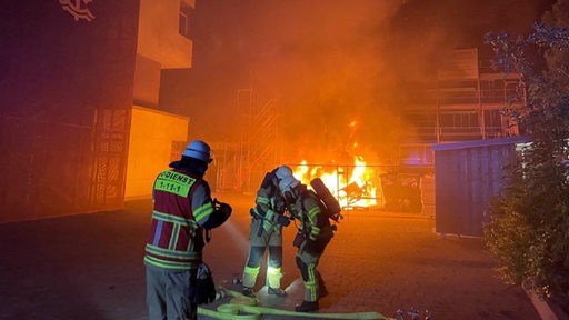 Mehrere Feuerwehrmänner ziehen einen Wasserschlauch über einen Schulhof, während im Hintergrund ein Feuer lodert.