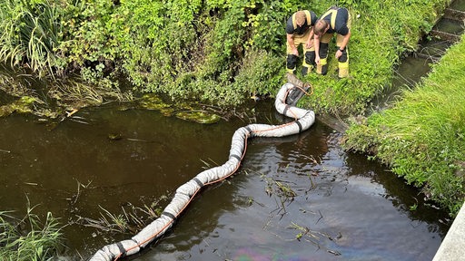 Feuerwehrmänner stehen neben einer Ölsperre am Flussufer.