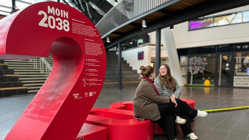 Zwei Personen sitzen auf einer roten Installation in einer Halle.