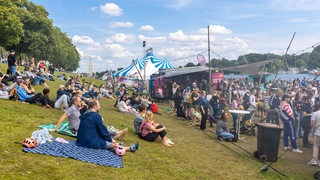 Menschen sitzen am Deich, Buden und Zelte sind an der Weser aufgebaut.