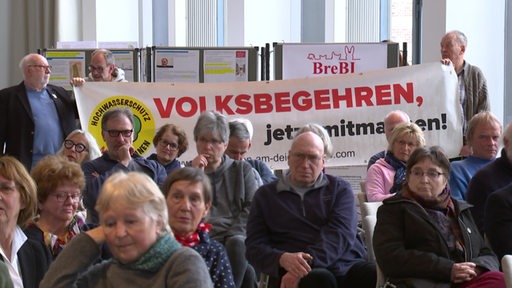 Personen sitzen in einem Raum, im Hintergrund stehen zwei Menschen, die ein Plakat mit der Aufschrift "Volksbegehren" hoch halten.