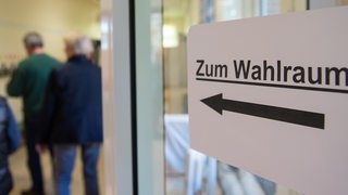 Menschen gehen in einer Grundschule an einem Hinweis mit der Aufschrift "Zum Wahlraum" vorbei,