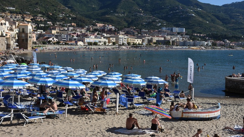 An einem Strand bei Cefalu auf Sizilien sitzen zahlreiche Touristen im Sand oder schwimmen im Wasser.