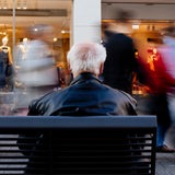 Ein Rentner sitzt auf einer Bank während vor ihm (mit Langzeitbelichtung) Menschen vorbeilaufen.