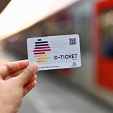 Ein Deutschlandticket wird vor einem Zug in die Kamera gehalten