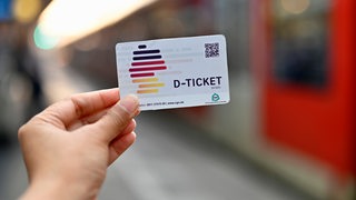 Ein Deutschlandticket wird vor einem Zug in die Kamera gehalten