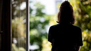 Eine Frau steht in ihrer Wohnung an einem Fenster.