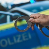 Ein Polizist steht vor einem Polizeiwagen und hält Handschellen in der Hand.