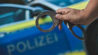 Ein Polizist steht vor einem Polizeiwagen und hält Handschellen in der Hand.