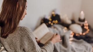 Eine junge Frau mit langen Haaren sitzt im Bett mit Wolldecke und liest in einem Buch.