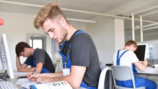 Drei junge Menschen sitzen in Blaumännern vor Büchern und Computern.