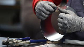 Zwei Hände in Schutzhandschuhen halten einen Metallring. Daneben liegt eine Zange.