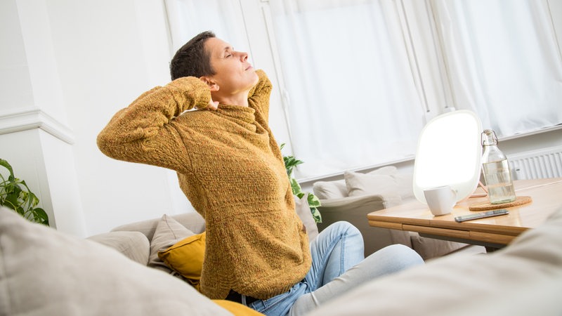 Eine Frau sitzt vor einer Tageslichtlampe und reckt sich (Symbolbild)