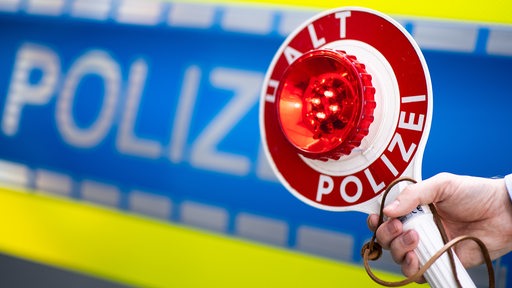 Eine Polizeikelle wird hochgehalten, im Hintergrund ein Polizeifahrzeug