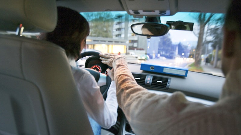 Ein Fahrlehrer greift einer Fahrschülerin ins Lenkrad.