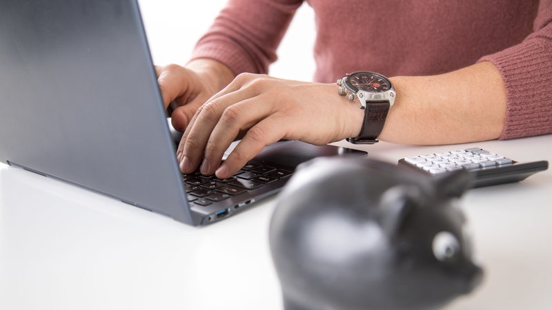 Ein junger Mann sitzt mit Taschenrechner und Sparschwein am Laptop.